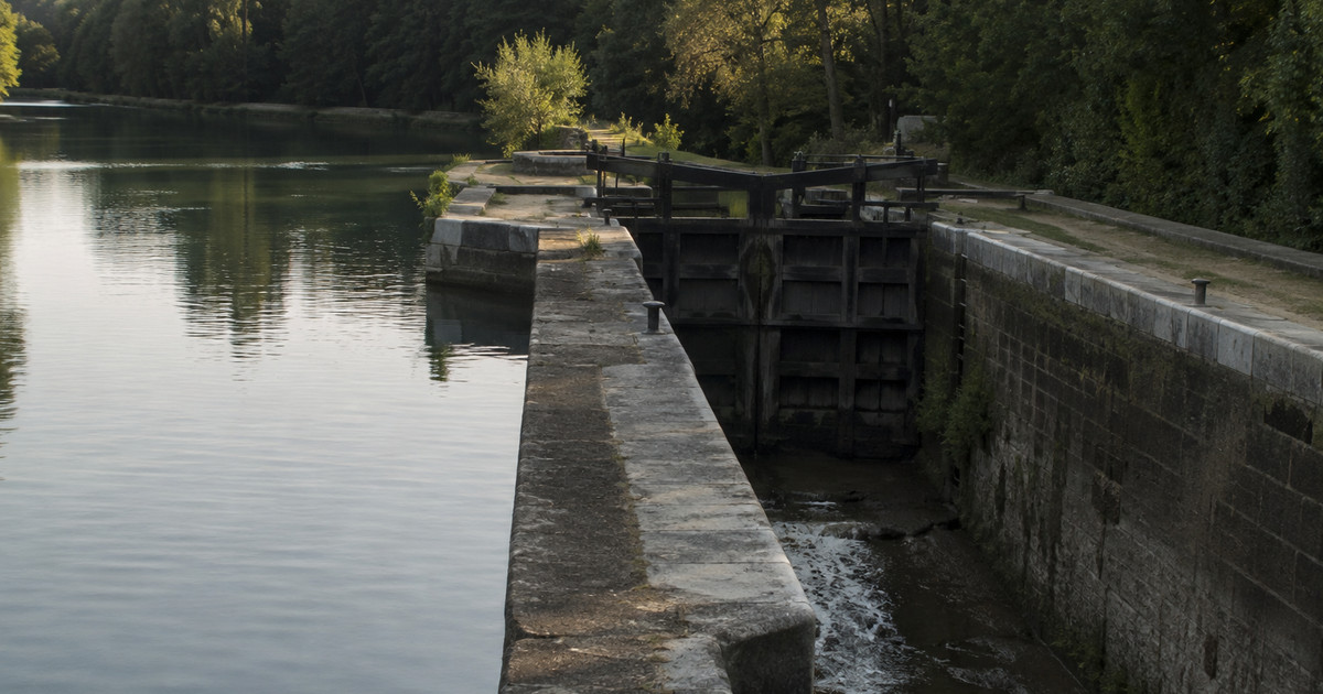 Écluse de canal vue depuis la berge : à gauche un bief plein et calme, au centre une porte fermée, à droite un bief presque vide où seul un filet d'eau s'écoule